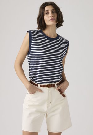 Vrouw met een mouwloos top met marineblauw-witte strepen, crèmekleurige shorts met hoge taille en een gevlochten bruine riem, staand met handen in de zakken.