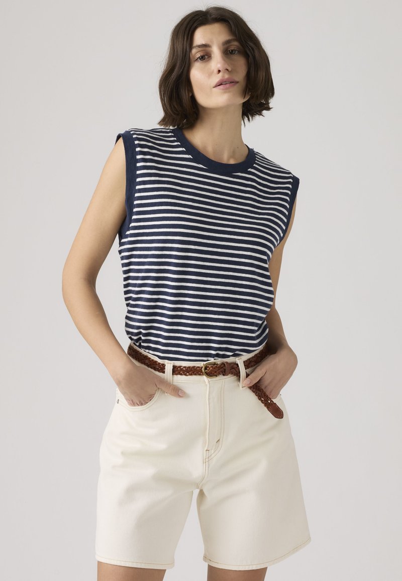 Γυναίκα φοράει navy και λευκή ριγέ αμάνικη μπλούζα, κρεμ ψηλόμεσα σορτς και πλεχτή καφέ ζώνη, στέκεται με τα χέρια στις τσέπες.