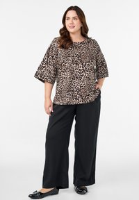 Naisel, kellel on lainelised pruunid juuksed, on seljas leopardimustriga pluus, laiad mustad püksid ja mustad lamedad kingad, millel on nailonist detailid, seistes valge taustaga.