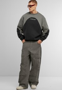 Sudadera de color bloqueado en gris oscuro y negro con puños acanalados y un logo, combinada con pantalones cargo gris claro que tienen varios bolsillos.