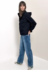 Femme portant une veste à capuche noire, un jean bleu à jambes larges et des baskets blanches à rayures vertes, debout de profil contre un fond uni.