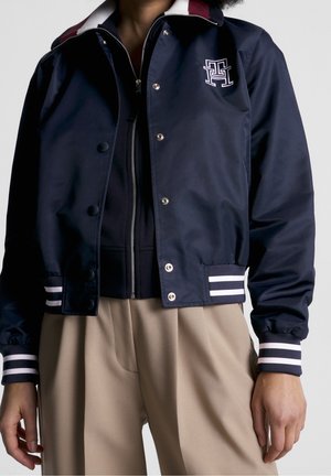 Person wearing a navy bomber jacket with white stripes on cuffs and hem and beige pants, jacket unzipped showing a black inner layer.