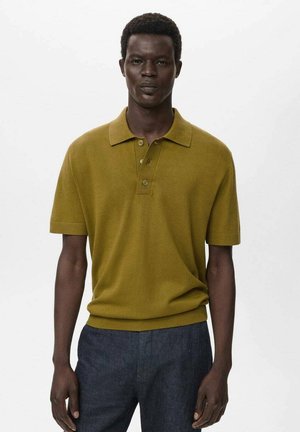 Khaki green polo shirt with a classic collar, short sleeves, three-button placket, and ribbed cuffs; paired with dark denim trousers.