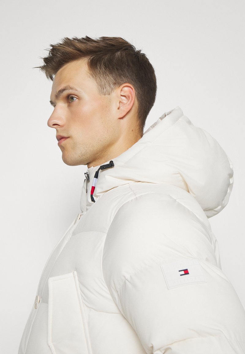 Chaqueta puffer blanca con capucha, con cierre de cremallera y bolsillos frontales. La manga izquierda tiene un parche con el logo. Textura suave y acolchada.