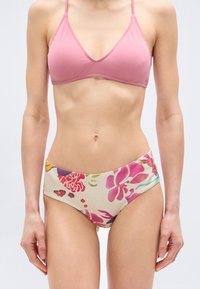 Γυναίκα που φοράει ροζ μπικίνι τοπ και φλοράλ μαγιό ψηλόμεσο, στέκεται με τα χέρια χαλαρά σε ένα απλό φόντο.