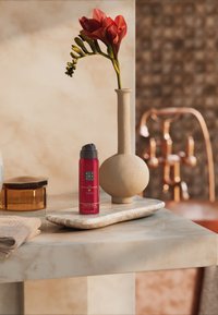 Spray rosso su un vassoio di marmo chiaro, accanto a un vaso di ceramica beige con fiori rossi, un contenitore dorato e un asciugamano morbido marrone.