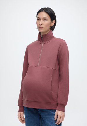 Gravid person iført en fiolett genser med halv glidelås og blå jeans, stående mot en enkel, lys bakgrunn.