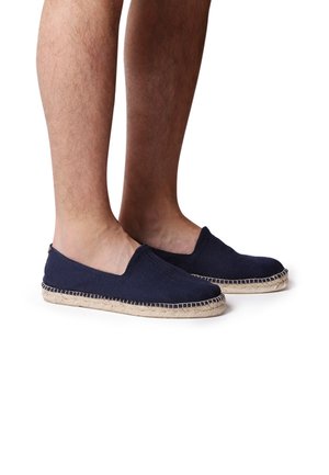 Jambes nues portant des espadrilles à enfiler bleu marine avec des semelles en jute tissé sur fond blanc.