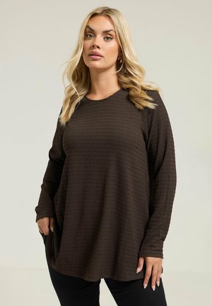 Blondynka w luźnej, ciemnobrązowej, długorękawowej teksturowanej bluzce i czarnych spodniach, stojąca na tle jednolitego jasnego tła.