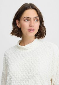 Weißer, strukturierter Pullover mit einem Cowl-Ausschnitt und einer gemusterten Oberfläche. Das Model hat kurze Haare und trägt kleine Creolen.
