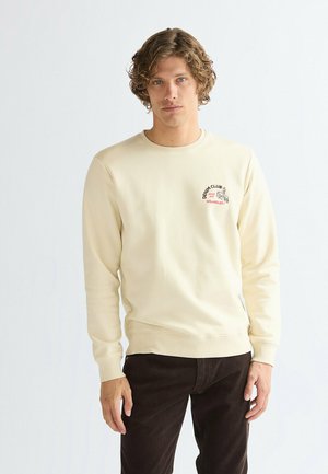 Jeune homme aux cheveux bouclés portant un sweat crème avec le texte « Denim Club Wrangler » et un pantalon foncé sur un fond clair uni.