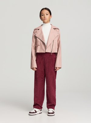 Giacca corta in ecopelle rosa con zip frontale, abbinata a pantaloni bordeaux ampi con trama e sneakers bianche con dettagli scuri.