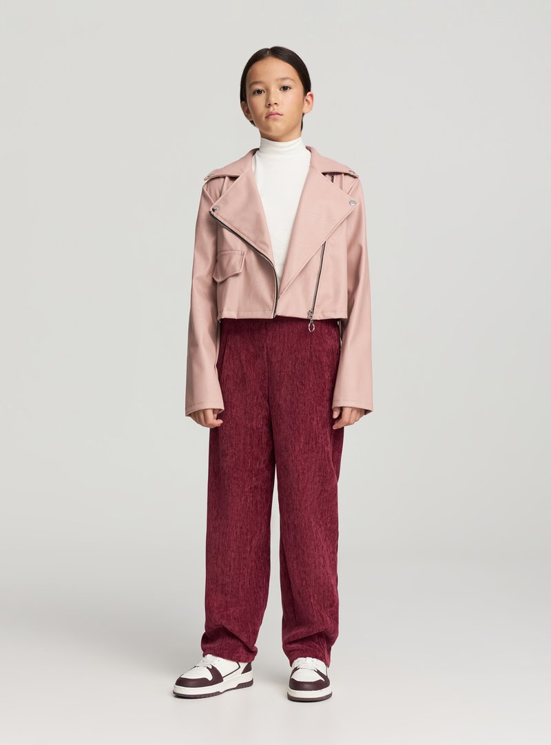 Giacca corta in ecopelle rosa con zip frontale, abbinata a pantaloni bordeaux ampi con trama e sneakers bianche con dettagli scuri.