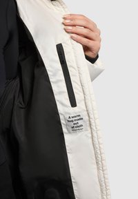 Vnútro bielej a čiernej prešívanej bundy so zipsovou kapsou a štítkom s nápisom „Teplé objatie z látky.“ Textúrovaná tkanina.