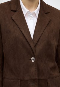 Gros plan d'une personne portant un blazer en daim marron par-dessus une chemise blanche boutonnée avec un seul bouton visible fermé.