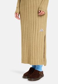 Beiges, geripptes Strickkleid mit langen Ärmeln und seitlichen Schlitzen, ausgestattet mit einem kleinen Logo-Patch. Kombiniert mit braunen Wildlederclogs und blauen Socken.
