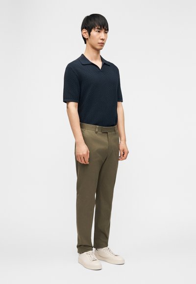 Άνδρας που στέκεται με τα χέρια χαλαρά, φοράει πλεκτό σκουρομπλε πόλο πουκάμισο, ελιά παντελόνια και λευκά αθλητικά παπούτσια μπροστά σε απλό λευκό φόντο.