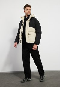 Chaqueta tipo puffer en negro y beige con capucha, cierre de cremallera, grandes bolsillos delanteros y una textura suave, combinada con pantalones negros y zapatillas.