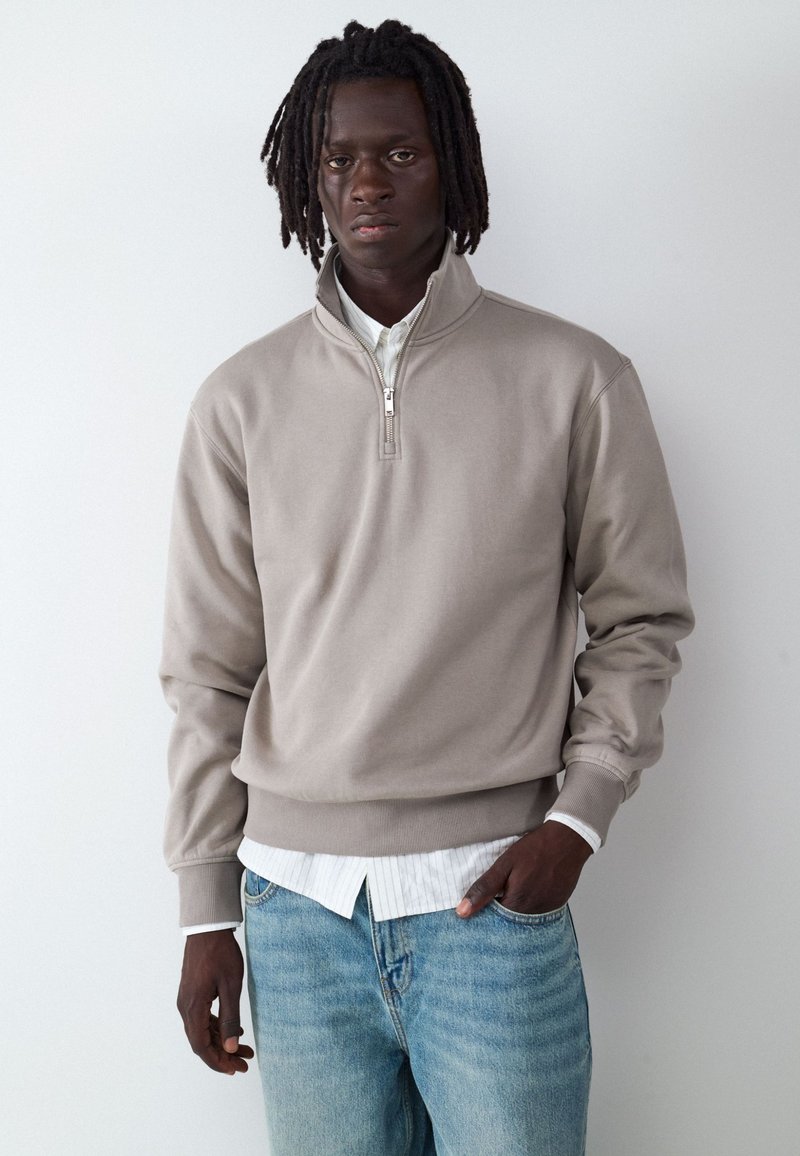 Jeune homme à la peau foncée et aux dreadlocks portant un sweat-shirt beige à fermeture éclair quart de zip sur une chemise blanche et un jean bleu clair, debout devant un fond blanc.