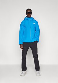 Blå vanntett jakke med hette, glidelås og logo på brystet, kombinert med svarte bukser og hvite sneakers. Matterte solbriller.