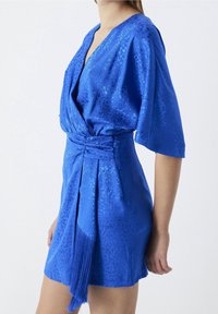 Robe en soie bleue avec une texture brillante, ourlet à franges courtes et manches de style kimono. Présente un design croisé et un détail de taille froncée.