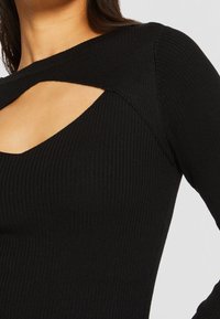 Svart ribbad stickad topp med en bred halsringning och geometrisk utskärning. Långa ärmar och en åtsittande design framhäver textur och struktur.