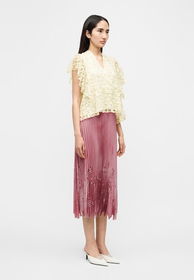 Τοπ από δαντέλα σε απαλό κίτρινο με στρωτές βολάν, συνδυασμένο με ροζ πλισέ φούστα με κεντήματα λουλουδιών και λευκές mule με τακούνι.