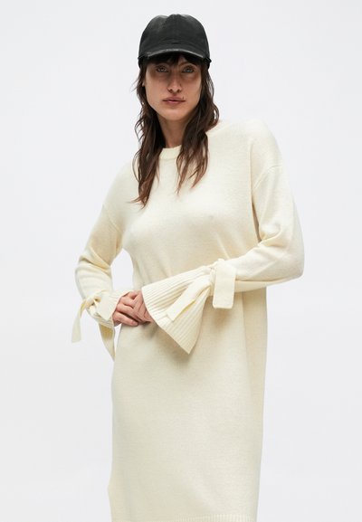 Πλεκτό κρεμ φόρεμα πουλόβερ με μανίκια καμπάνα και κορδόνια. Απαλή υφή και άνετη εφαρμογή, με στρογγυλή λαιμόκοψη.