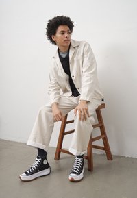 Svetle béžová plátěná bunda, černá košile, krémové kalhoty a černé platformové tenisky. Pozadí se dřevěným stoličkem; minimalistické studio.
