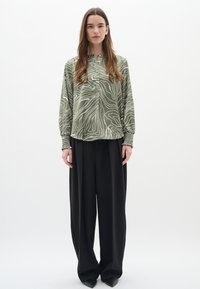 Blusa verde oliva con linee ondulate bianche, colletto alla cinese e polsini elasticizzati, abbinata a pantaloni larghi neri e scarpe a punta.
