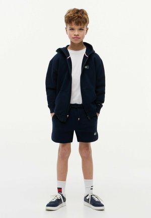 Garçon debout face à l'avant portant un sweat à capuche zippé bleu marine, un t-shirt blanc, un short bleu marine, des chaussettes blanches à rayures et des baskets bleu marine avec des lacets blancs.