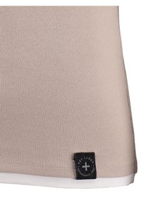 Hellbeige, gerippter Tanktop mit weißem Saum, ausgestattet mit einem schwarzen Logoetikett am unteren Ende. Glatte Textur mit tailliertem Design.
