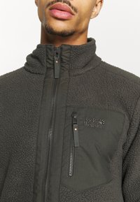 Mężczyzna w ciemnoszarej polarowej kurtce z zamkiem błyskawicznym z przodu oraz zamkniętą kieszenią na piersi, wyhaftowaną logo "Jack Wolfskin".