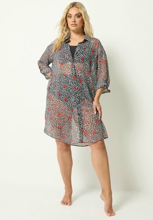 Femme blonde portant une robe transparente à imprimé léopard avec des motifs de cerises rouges, debout pieds nus sur un fond blanc uni.