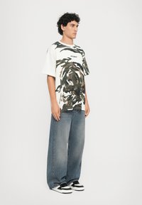 White cotton t-shirt featuring a green and brown tiger graphic, paired with loose-fitting blue denim jeans. Black and white sneakers complete the look.