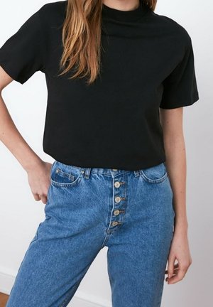 Woman wearing a black short-sleeve shirt tucked into high-waisted blue jeans with multiple visible front buttons.