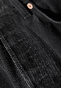 Tejido de mezclilla negro con una superficie ligeramente texturizada. Presenta costuras visibles y un acento de botón de cobre cerca de la parte superior.