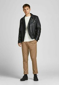 Giacca da biker nera in pelle sopra un maglione bianco a coste, abbinata a pantaloni beige e scarpe nere con lacci. Texture lisce e design audace.