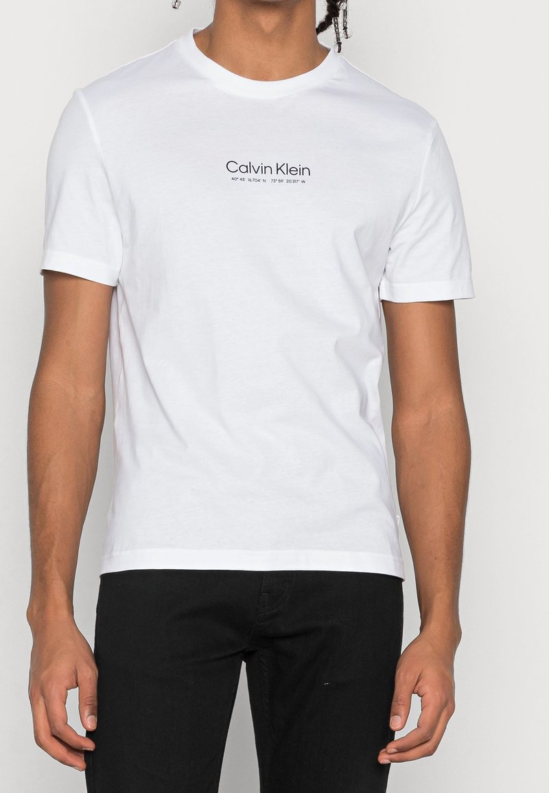 T-shirt en coton blanc à manches courtes et col rond. Présente le logo texte "Calvin Klein" sur la poitrine en bleu marine, design simple.