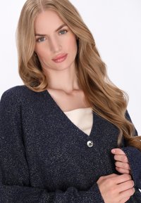 Granatowy kardigan z delikatnym połyskiem, z długimi rękawami, dekoltem w serek i jednym okrągłym srebrnym guzikiem. Tkana tkanina o fakturze.