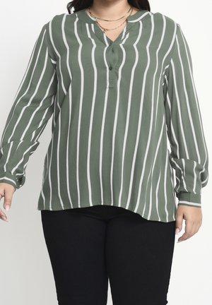 Femme portant un chemisier vert à manches longues avec des rayures blanches verticales, associé à un pantalon noir et des colliers dorés superposés.