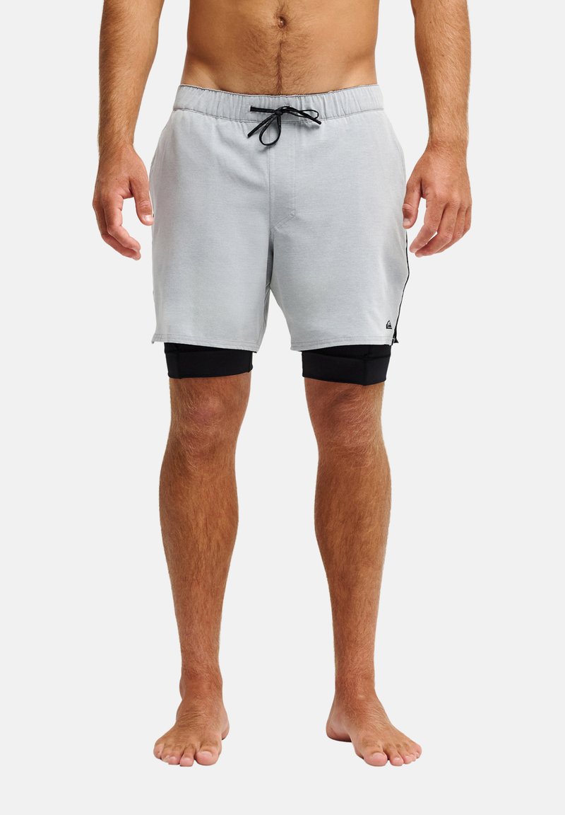 Homme portant un short gris clair à cordon par-dessus un short de compression noir, debout pieds nus devant un fond blanc.