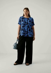 Top azul com padrão e mangas curtas, adornado com formas abstratas, combinado com calças largas pretas. Acessorizado com uma pequena bolsa azul e sandálias.