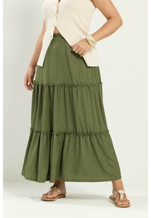 Kvinde iført en lagdelt maxinederdel i olivengrøn, cremet ærmeløs knapbluse, beige sandaler og et marmoreret armbånd om håndleddet.