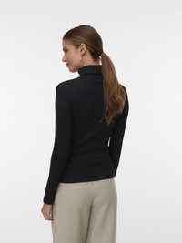 Hosszú barna lófarokkal rendelkező nő fekete garbós pulóverben és bézs nadrágban, az oldalára fordulva áll egy fehér háttér előtt.