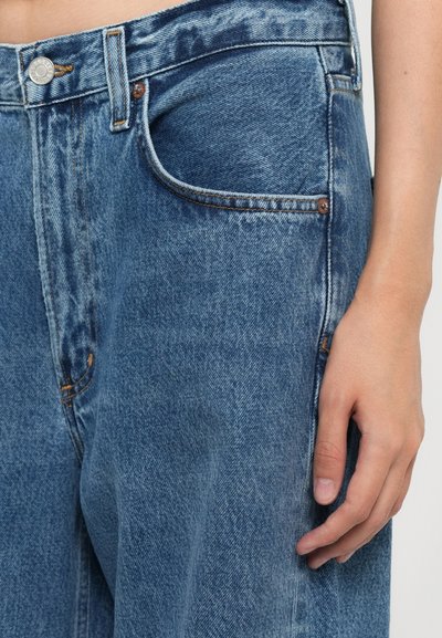 I jeans in denim azzurro chiaro presentano una vestibilità rilassata, cinque tasche e cuciture arancioni. Il tessuto mostra una leggera texture e dettagli di usura.