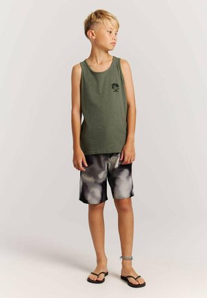 Blond jongetje met een groen hemdje, zwarte en grijze tie-dye korte broek, zwarte teenslippers en een blauwe enkelband, staand en kijkend naar rechts.