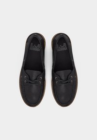 Ζευγάρι μαύρα δερμάτινα μοκασίνια με καφέ λαστιχένιες σόλες και κορδόνια, σε θέα από πάνω πάνω σε απλό φόντο.