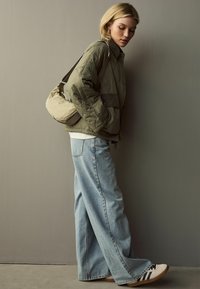 Olivgrüne gesteppte Jacke, weißes Oberteil, hellblaue weit geschnittene Jeans, beigetägige Umhängetasche und beige Schuhe mit schwarzen Streifen.