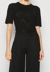 Kvinna i en svart kortärmad topp med diamantmönster och högmidjade svarta byxor, stående mot en enfärgad bakgrund.